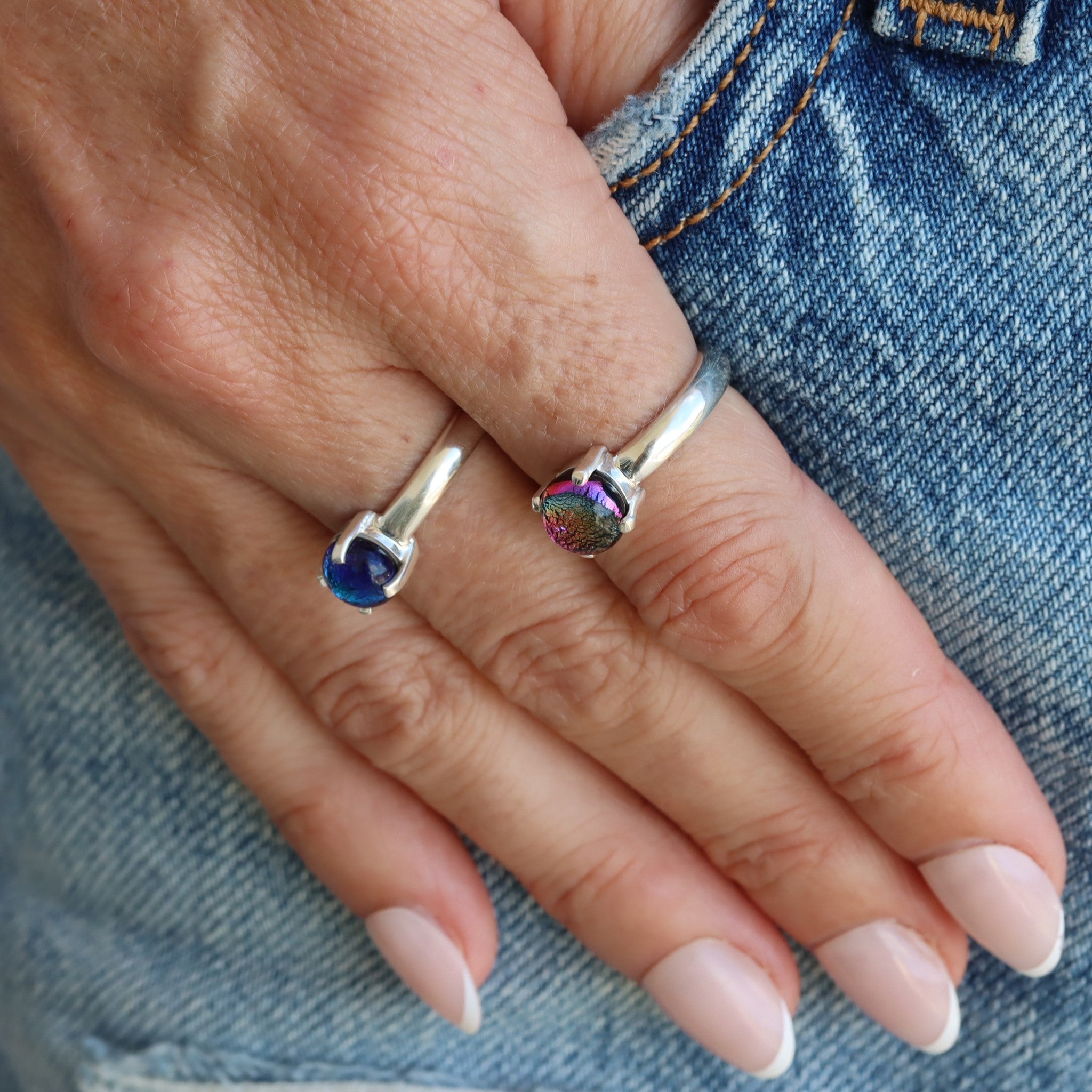 A hand wearing two sterling silver, small, round, claw-set dichroic glass gem rings.  One in pink and the other in blue tones.