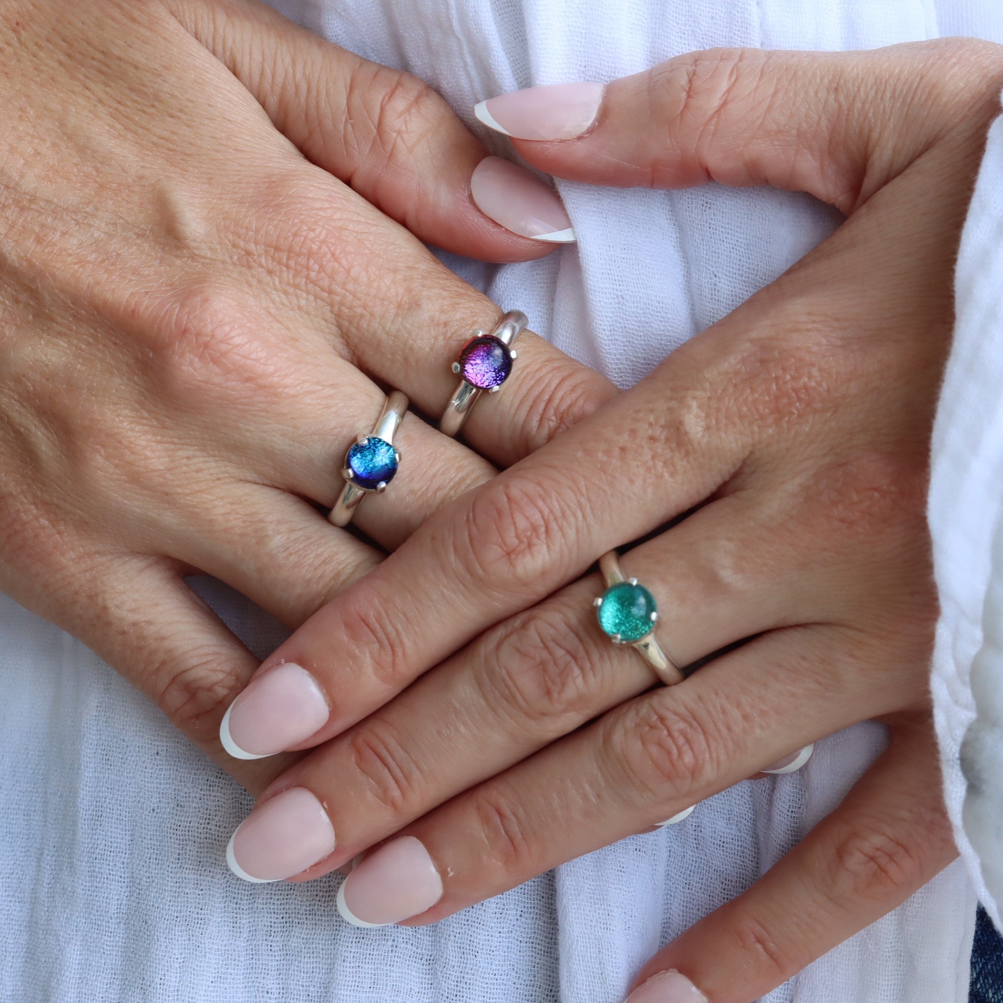 A model's hand wearing three sterling silver claw-set small round dichroic glass gem rings in pink, blue, and turquoise tones.