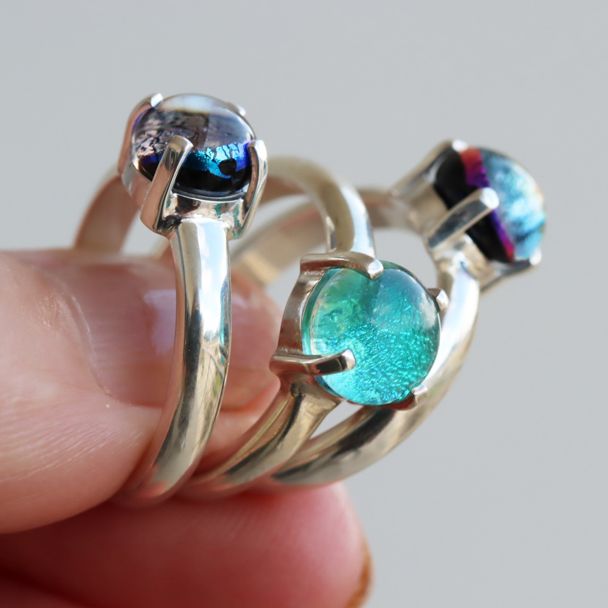 Fingers holding three sterling silver, round, small claw-set dichroic glass gem rings.  One in pink, another in blue and the other in turquoise tones.