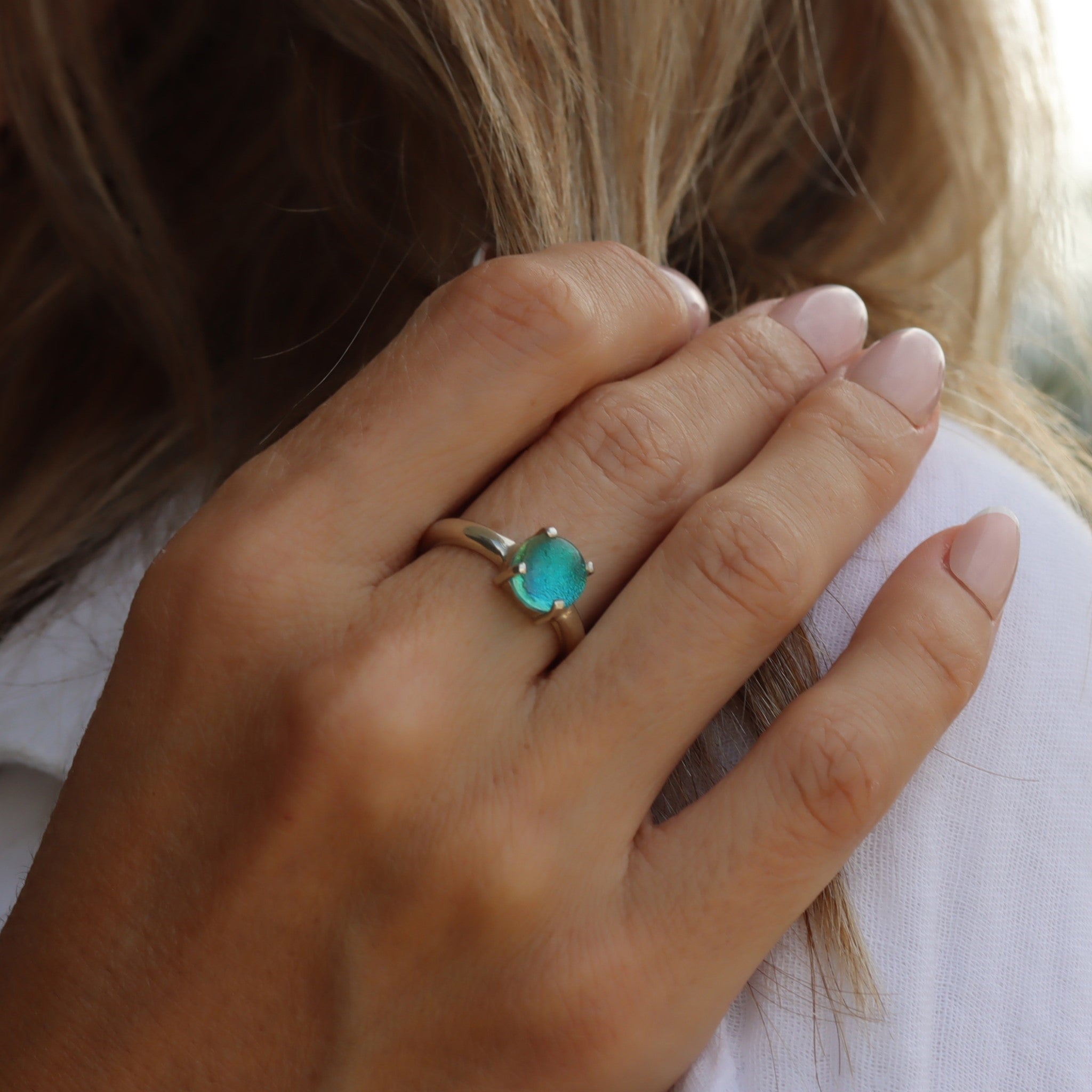 A model's hand wearing a sterling silver round, small, claw-set dichroic glass gem ring in turquoise tones.
