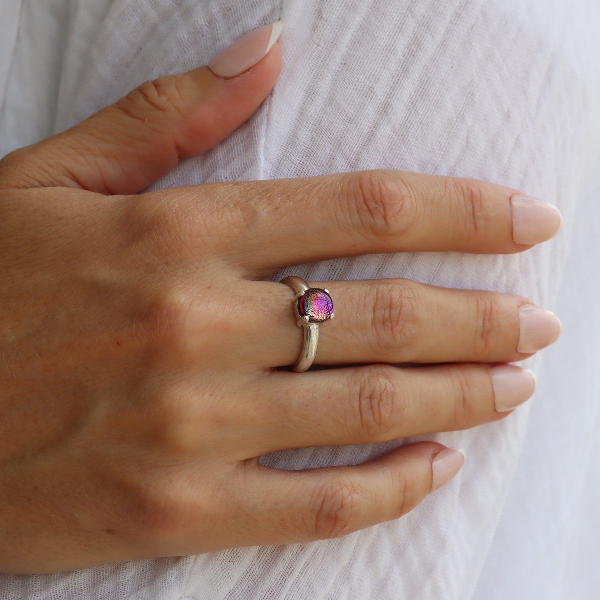 Model wearing a sterling silver round, small, claw-set dichroic glass gem ring in pink tones.